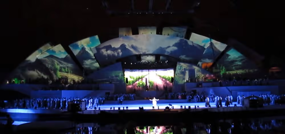 Panoramic scene with mountain landscape projected onto the scenography and performers on stage.